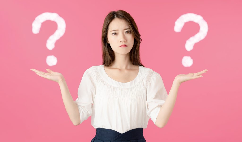 ピンクの背景の前で、女性が両手を広げて困った表情をしており、左右にクエスチョンマークが浮かんでいる