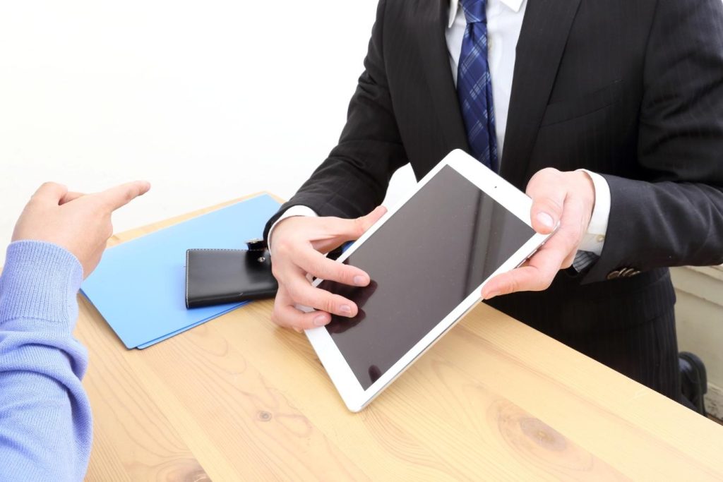 机に向かい合わせてタブレットを見せながら説明するスーツの男性と画面を指さす人