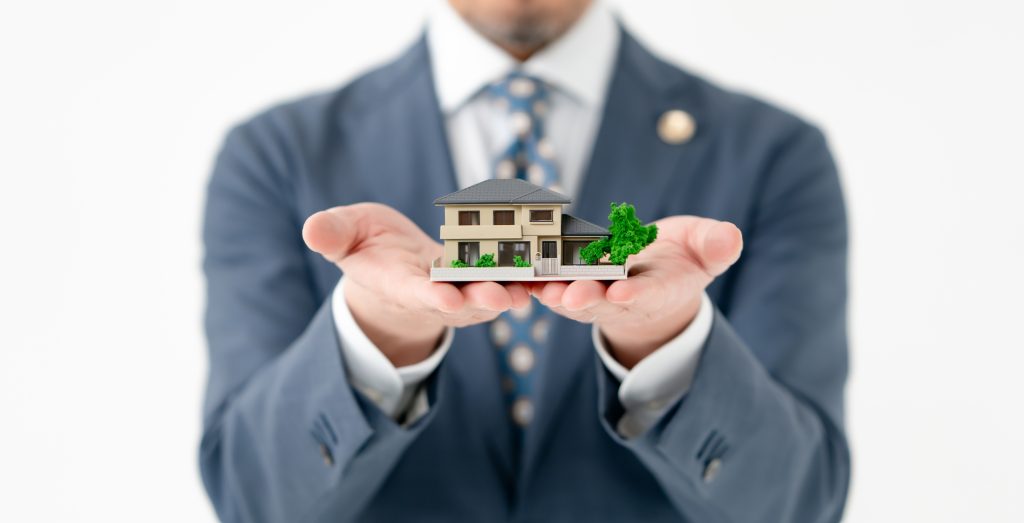 両手で一戸建ての住宅模型を差し出す、スーツ姿の男性