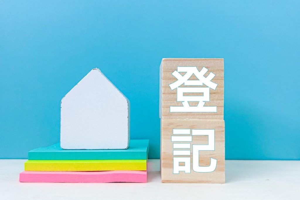 積まれた本の上に置かれた家の模型と「登記」と書かれた木のブロック