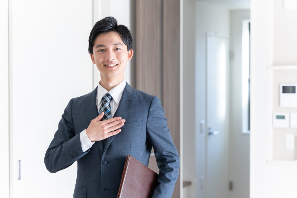 スーツ姿の男性が室内で微笑みながら胸に手を当て、書類ファイルを抱えている