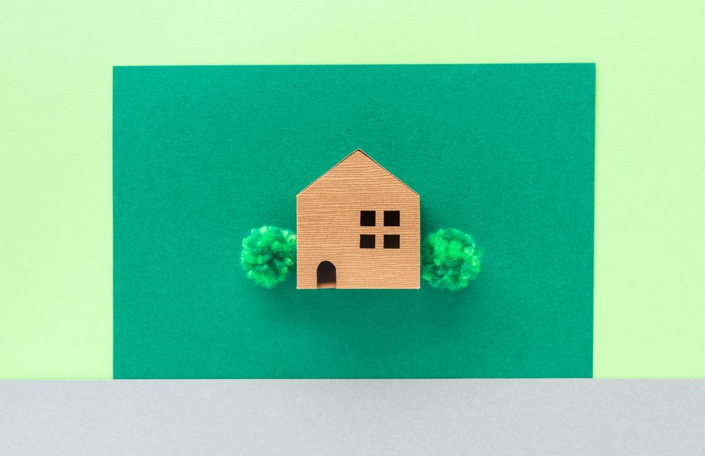 緑の背景の中央に、シンプルな木製の家の模型が置かれ、左右に小さな木のオブジェが添えられている