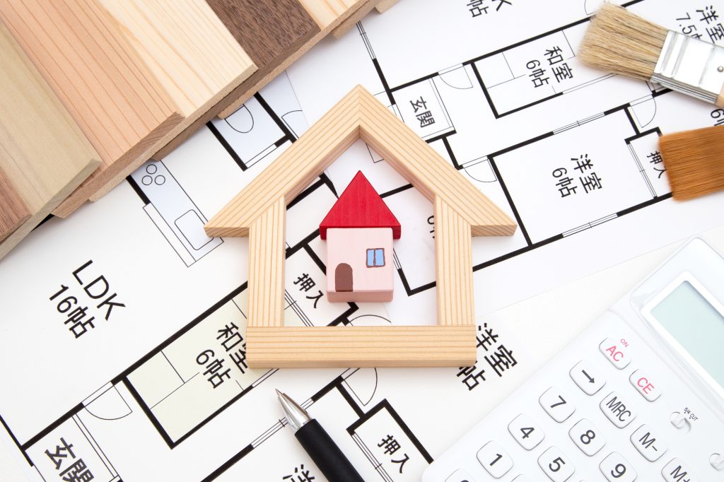 木材で家型に囲まれた住宅模型が、間取り図、電卓、ペン、木材サンプルの上に配置されている写真