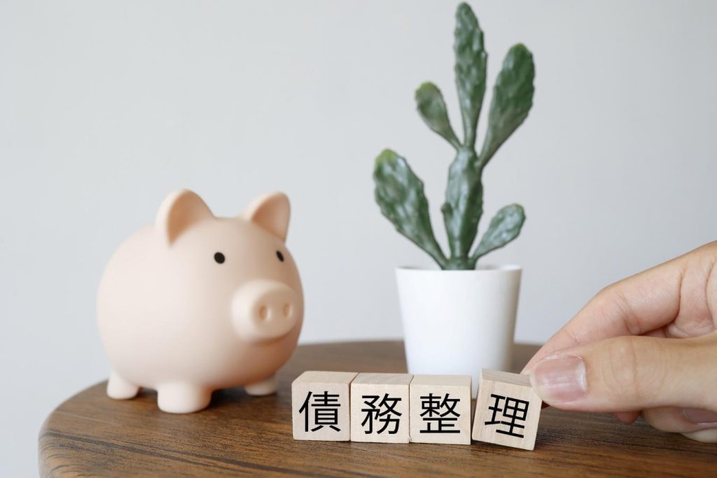 「債務整理」と書かれた木のブロックとブタの貯金箱、観葉植物