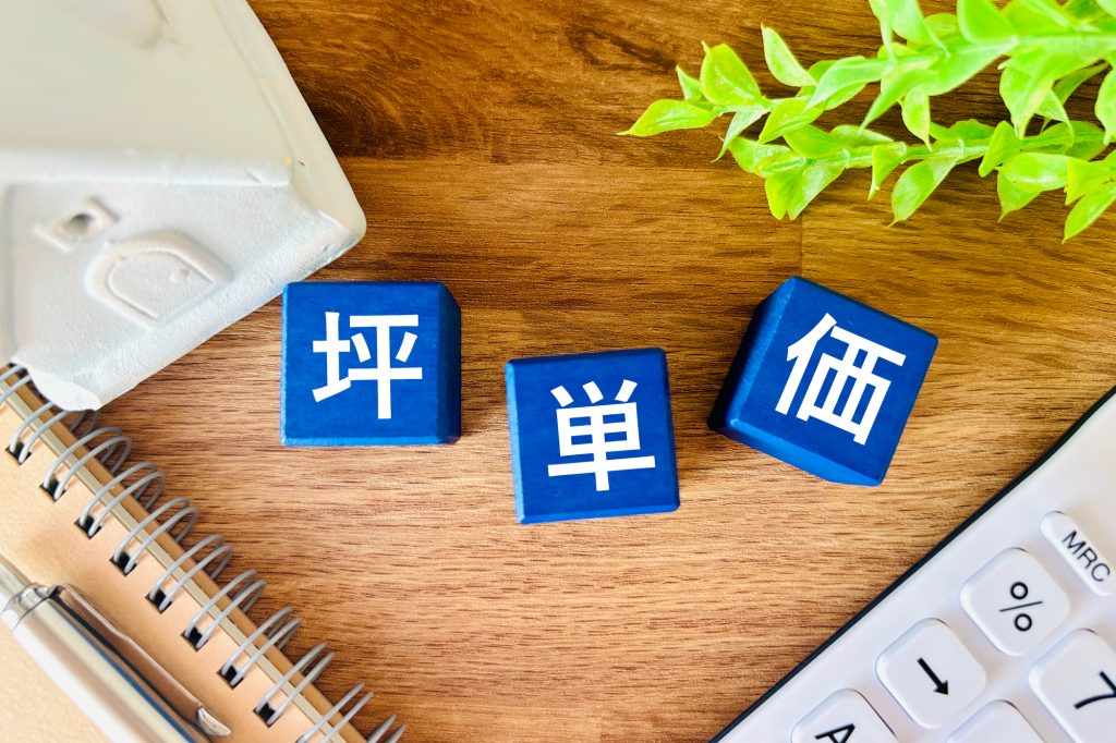 青い立方体に「坪単価」の文字が並び、ノート、ペン、電卓、家模型の一部が配置されている画像