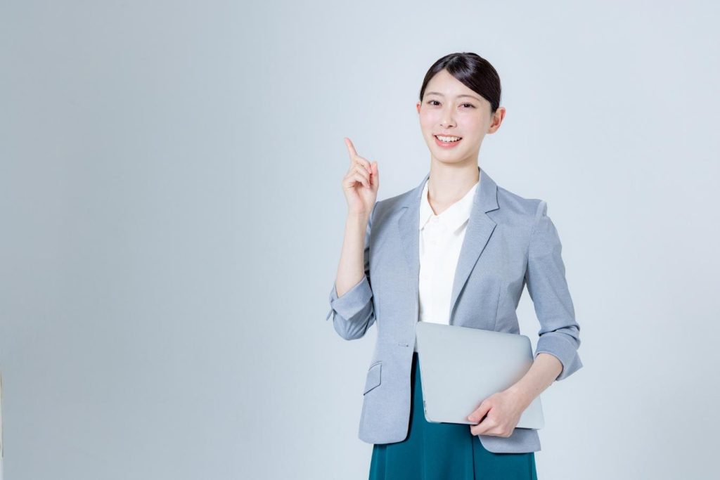 ノートパソコンを持って、もう一方の手の人さし指を立てたオフィスカジュアルの女性
