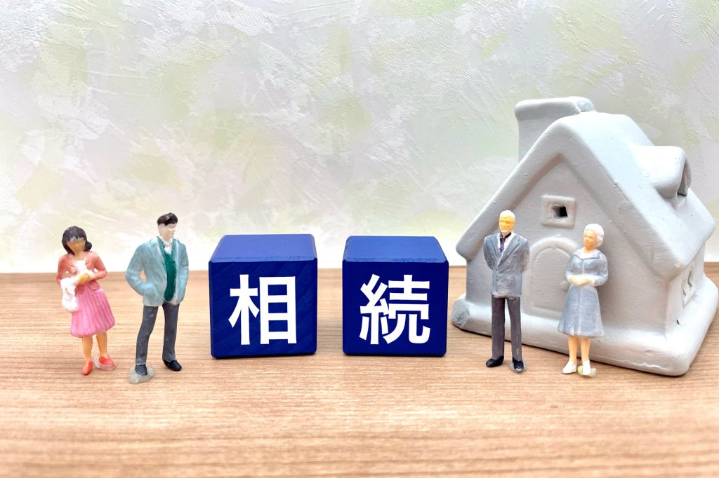 「相続」と書かれた青いブロックを挟んで並ぶ、家族のミニチュアフィギュアと家の模型