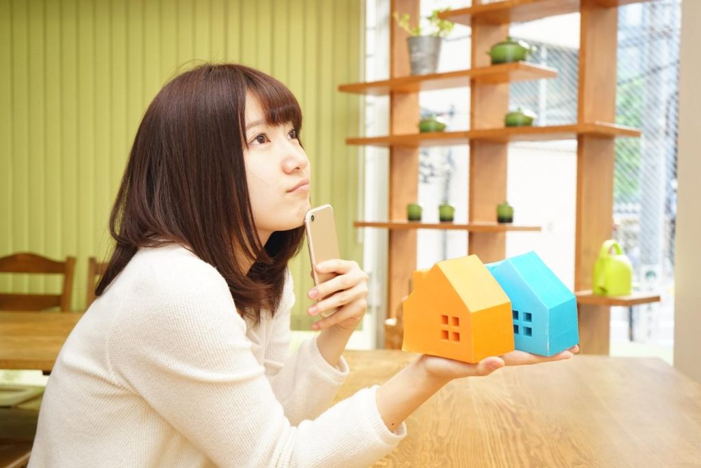 スマートフォンと2つの家の模型を持って考え込む女性