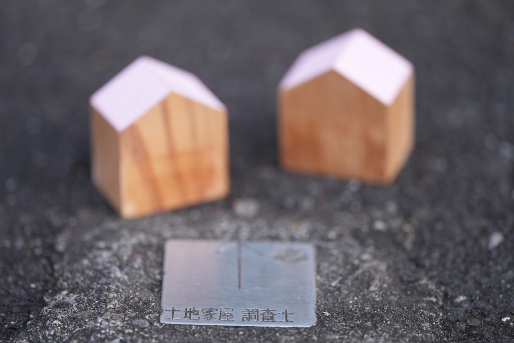 アスファルトに設置された「土地家屋調査士」の境界プレートと、奥に並ぶ2つの木製ハウスミニチュア