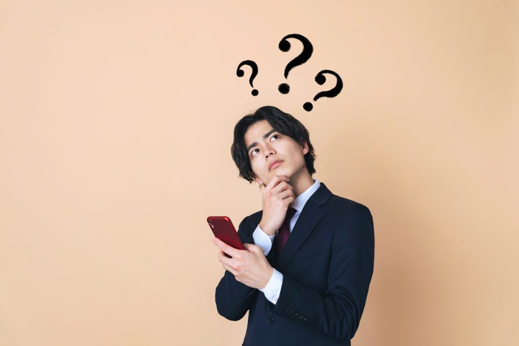 スマートフォンを片手に、頭上にクエスチョンマークを浮かべて考え込むスーツ姿の男性