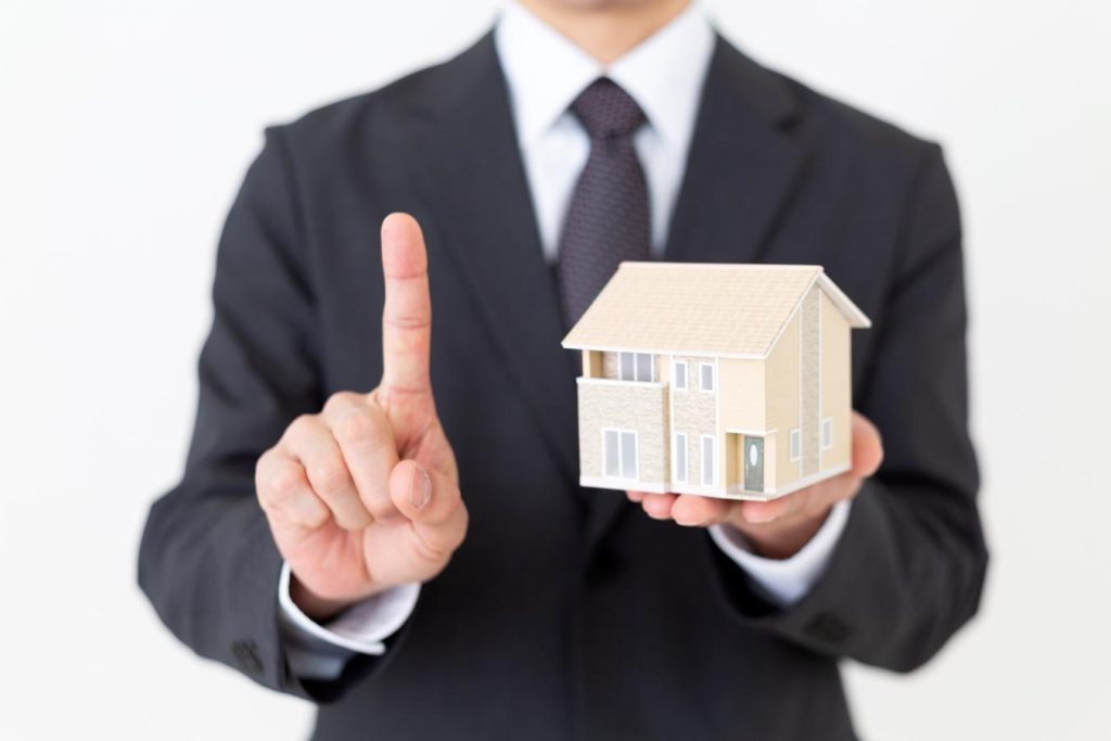 一戸建ての住宅模型を持って人差し指を立てるスーツの男性