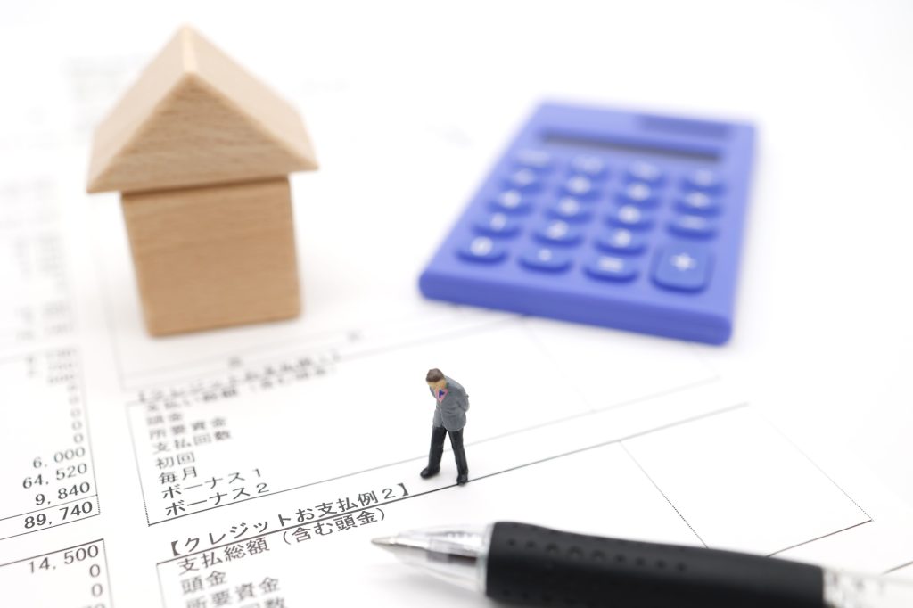 住宅ローンの支払明細書の上に置かれた家型オブジェ、電卓、ペンと考え込む人物模型