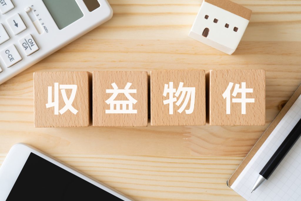 「収益物件」と書かれた木製ブロックと電卓、スマートフォン、住宅模型