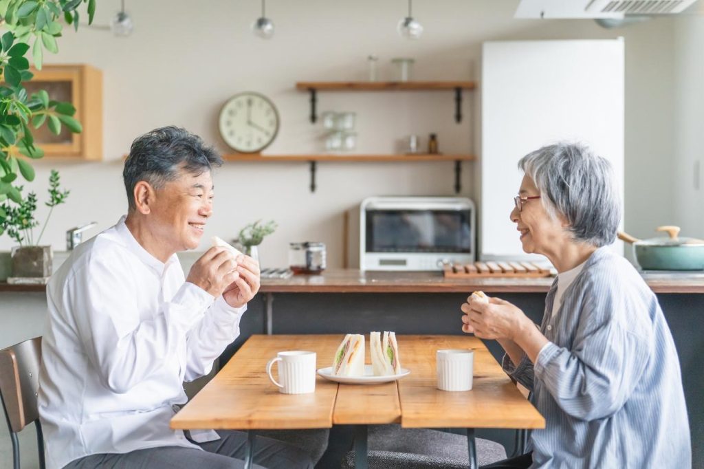ダイニングテーブルで向かい合い、笑顔でサンドイッチを食べるシニア夫婦
