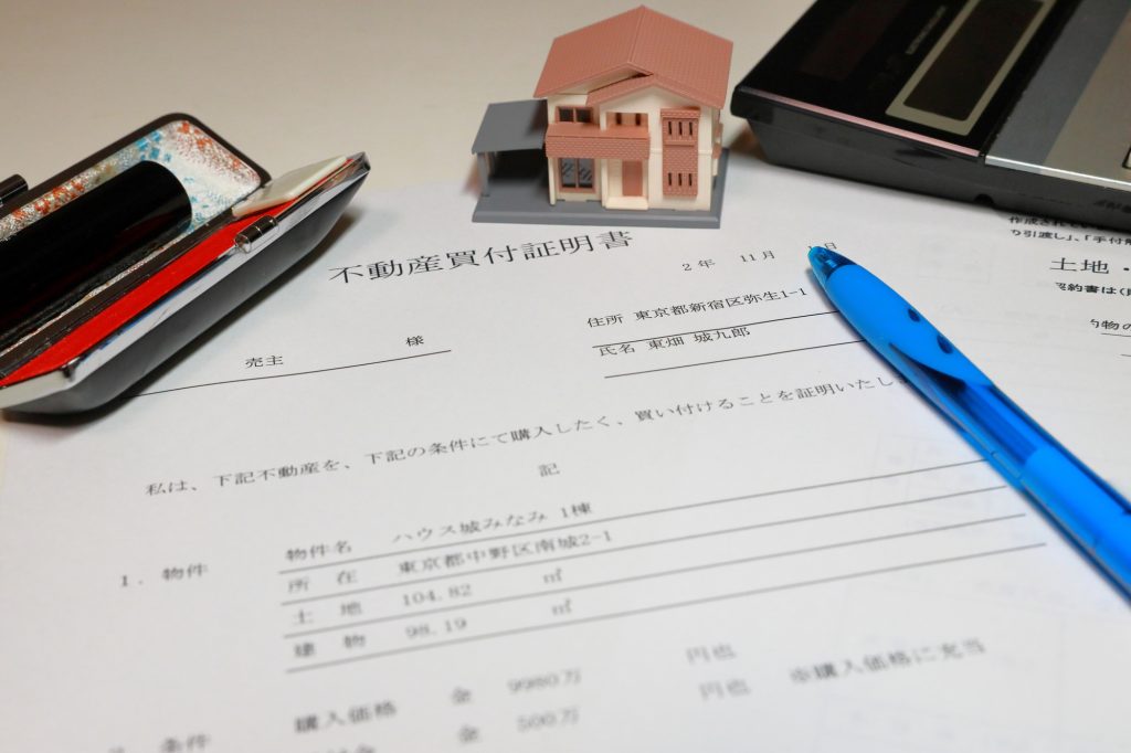 デスクに置かれた「不動産買付証明書」の書類、青いペン、赤い屋根の家の模型